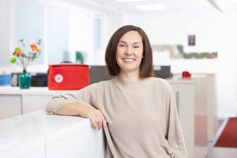 A woman stands in a hallway and smiles.